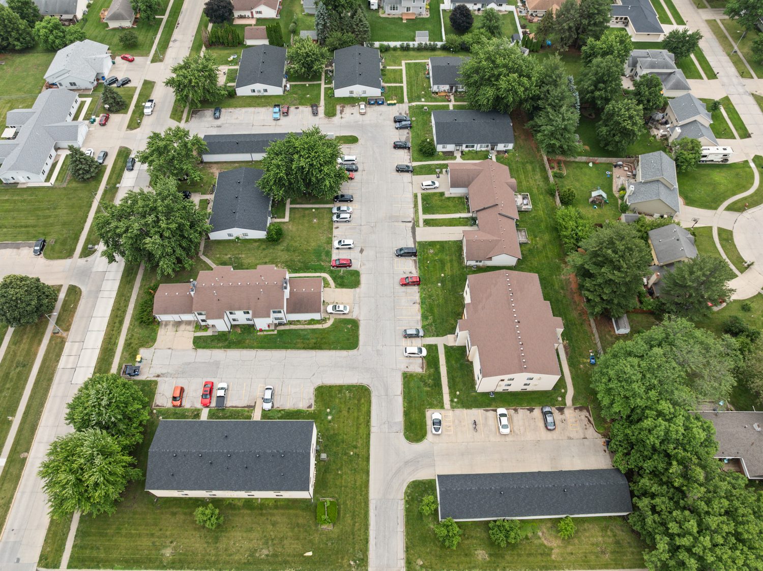 Aerial view straight down on East Park: five single-story fourplexes with dark roofs along the drive, the townhomes and apartment buildings with brown roofs, garages, parking and mature trees between the buildings.
