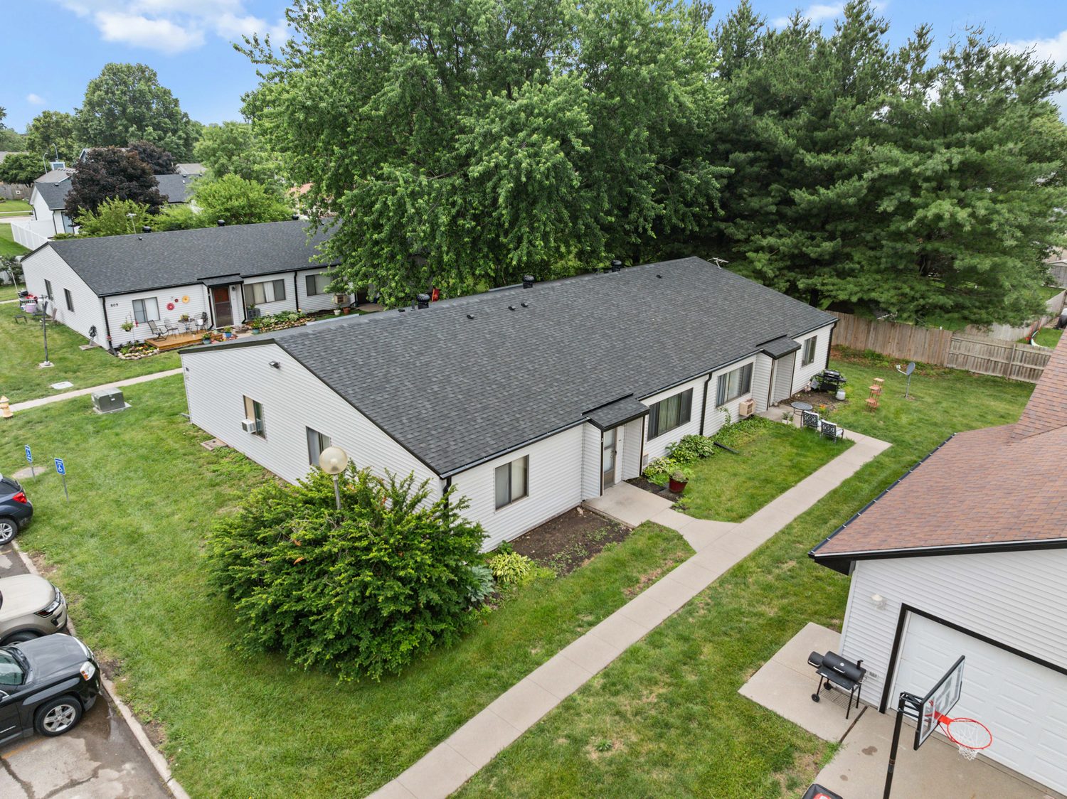 A single-story fourplex at East Park from above, new dark shingle roof, private front doors along the walk, and a flower bed and chairs on the lawn.