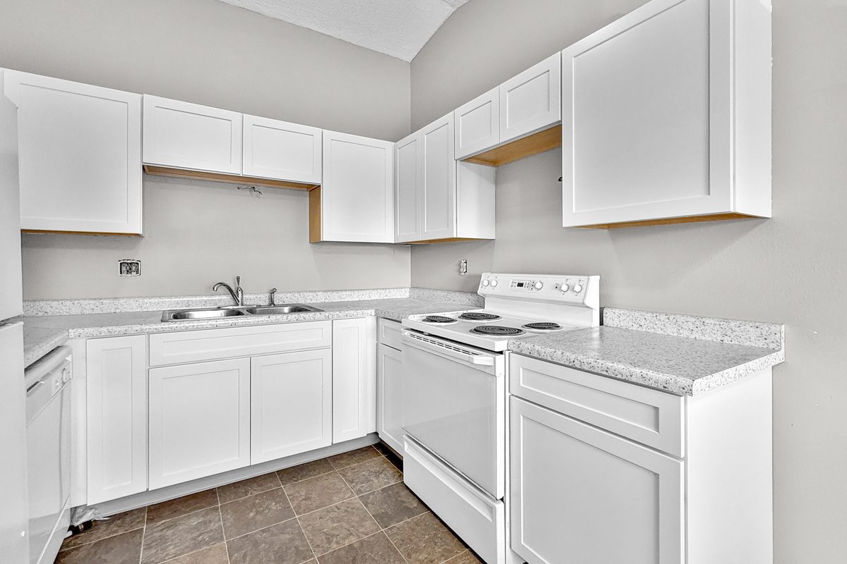 A renovated kitchen with white shaker cabinets, a grey countertop, a white range and a white dishwasher, open to the carpeted living room.