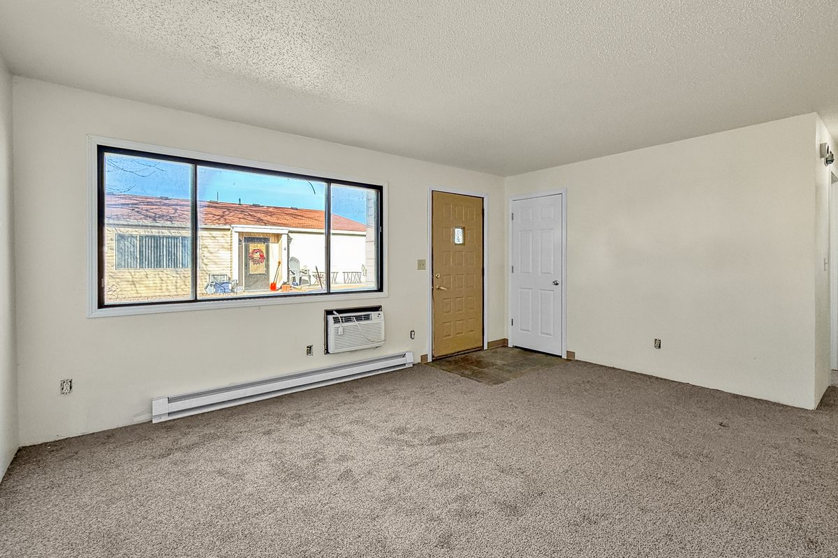 The living room of a fourplex apartment with tan carpet, white walls, a wide window, a wall air conditioner and the front door.