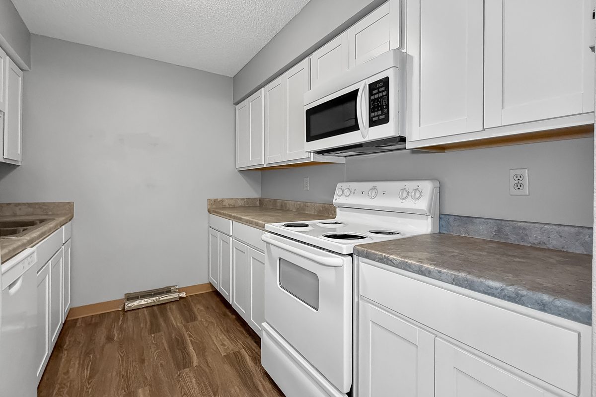 The kitchen of a renovated one bedroom apartment, white cabinets, a white range with an over-the-range microwave, and wood-look vinyl floor.
