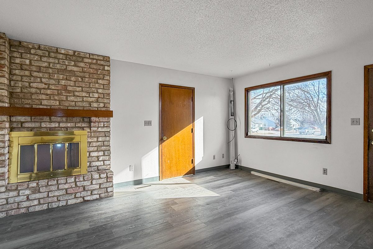 The living room of a townhome with a brick fireplace, wood-look plank floor, a window and the front door.