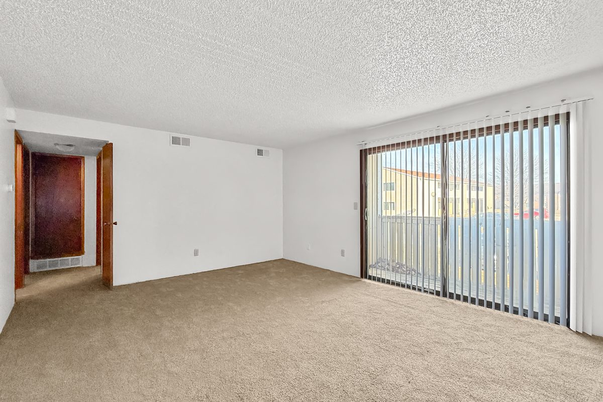 The living room of a two bedroom apartment in the twelve-unit building, carpeted, with a sliding glass door to the balcony and vertical blinds.