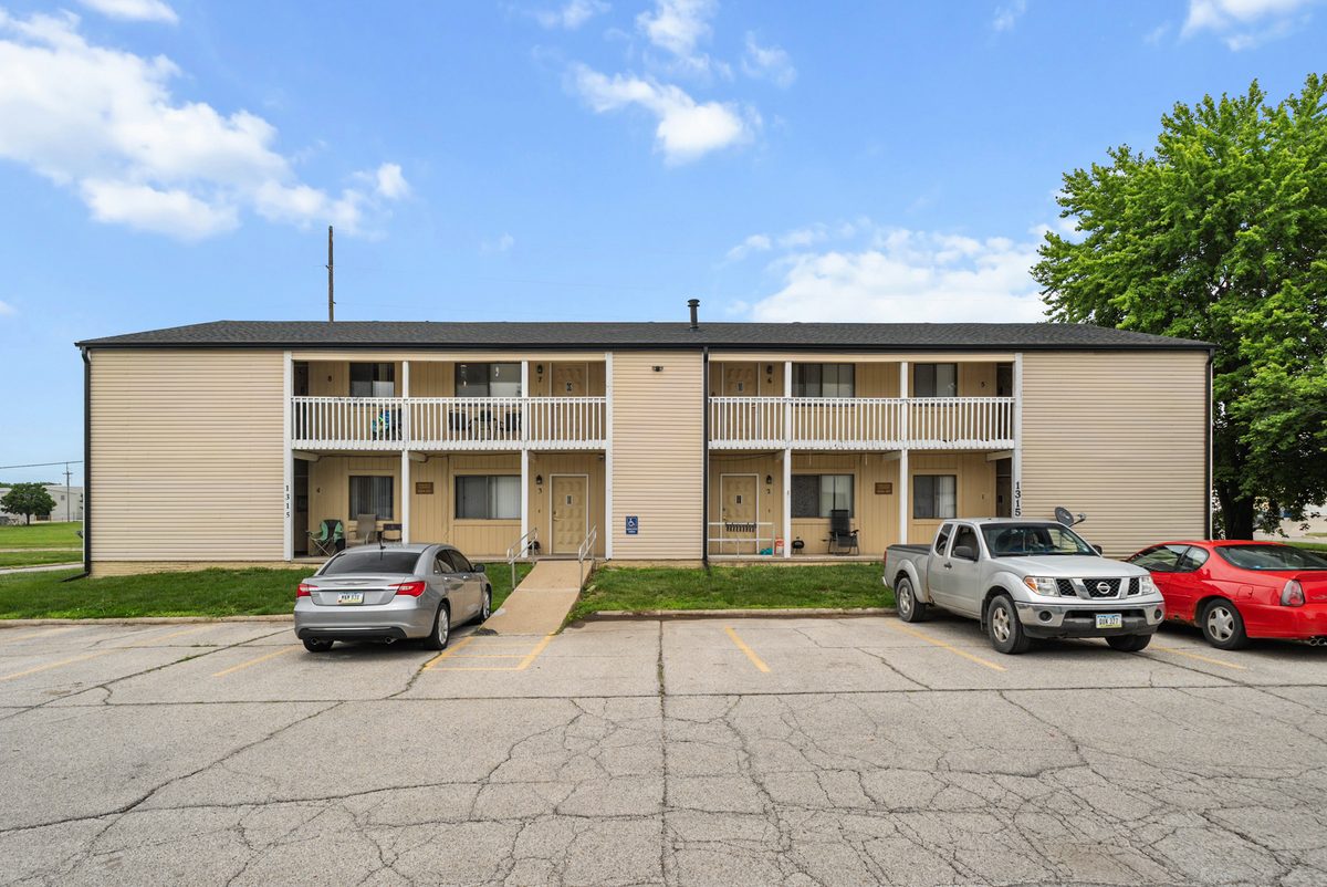 The front of the two-story eight-unit building, tan siding with a white balcony rail across the second floor and cars parked in front.