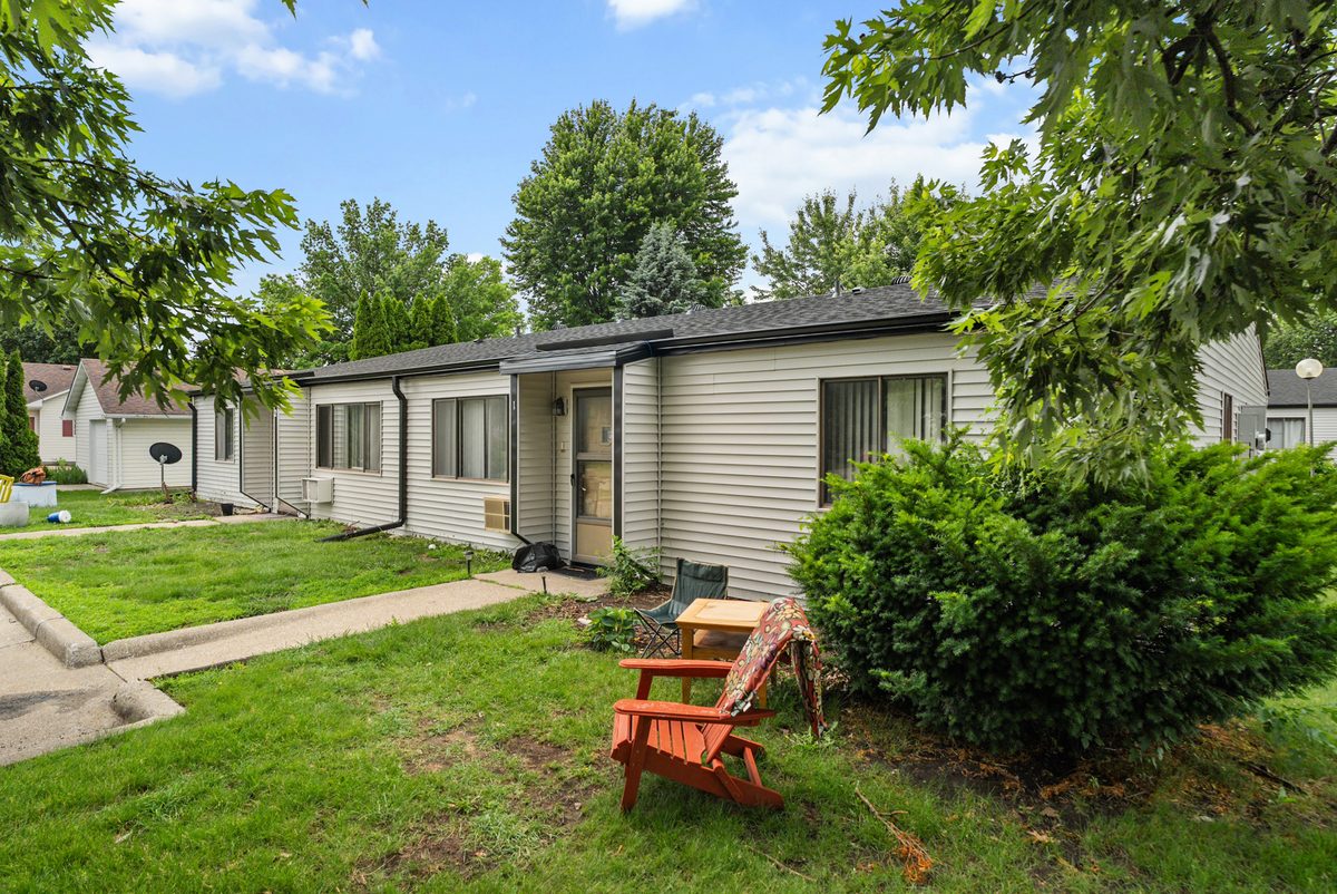 A single-story fourplex under mature trees, with a private front door and a red chair on the lawn.