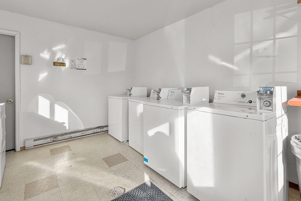 The community laundry room, with a row of white washers and dryers along the wall and windows above them.