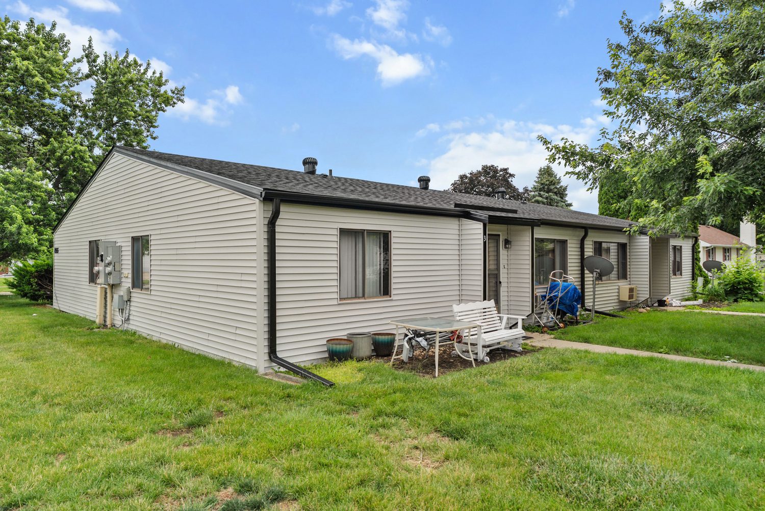 A single-story fourplex at East Park in summer, light siding with black trim, a new dark roof, private front doors along the walk and mature trees behind it.
