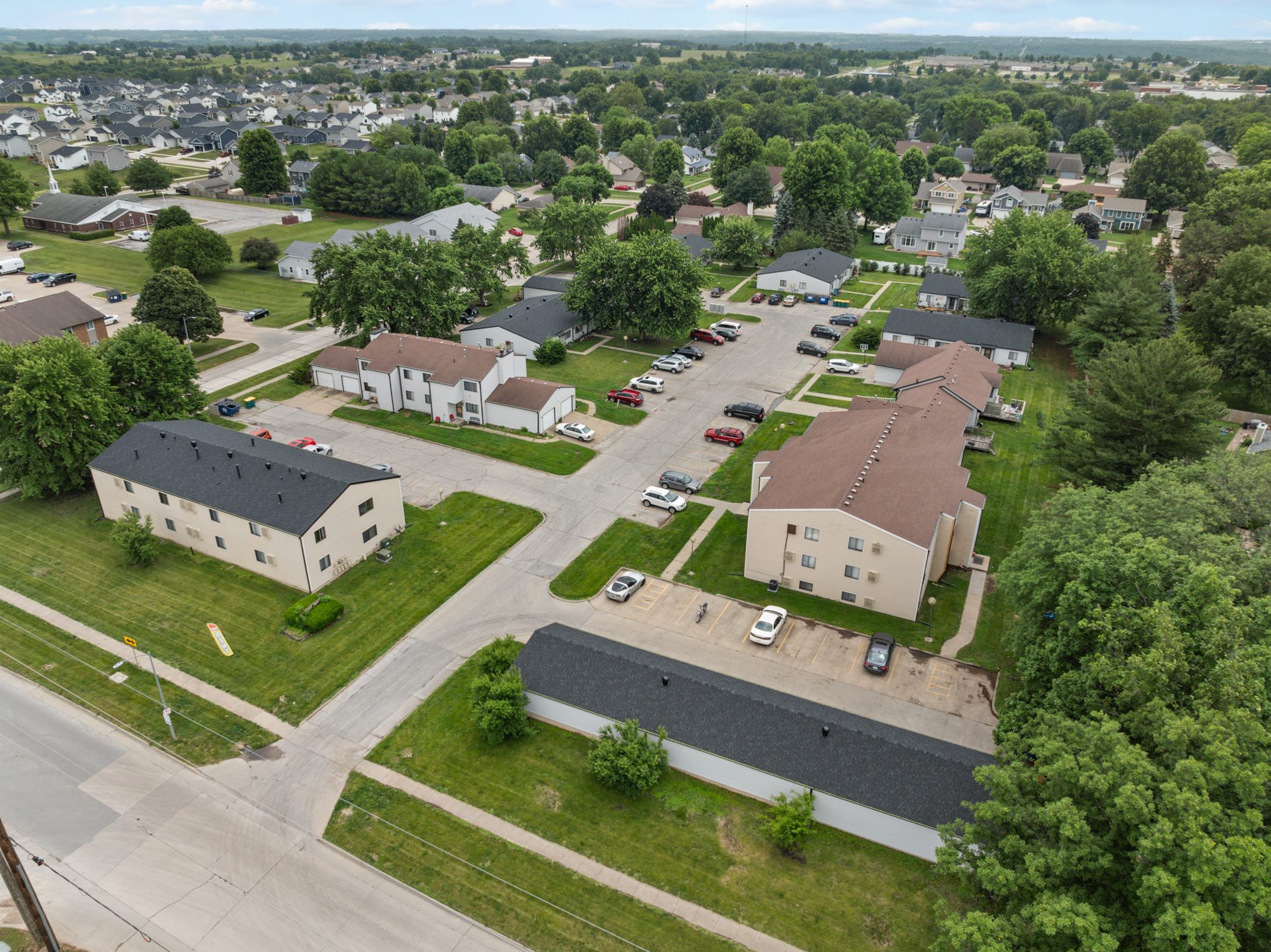 Aerial view of East Park Apartments in summer from the corner of N 14th Street: the two-story eight-unit building and a garage building in front, the three-story twelve-unit building at the right, the townhomes with attached garages behind them, and Indianola stretching to the horizon.